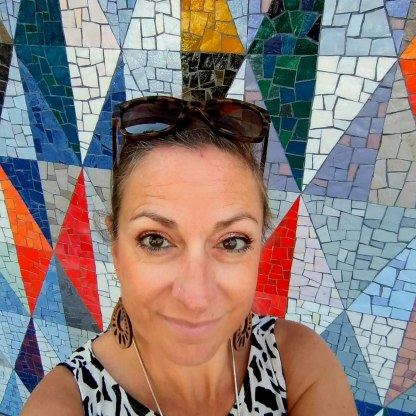 white woman with brown hair tied back, sunglasses and earrings in front of a colourful tiled background