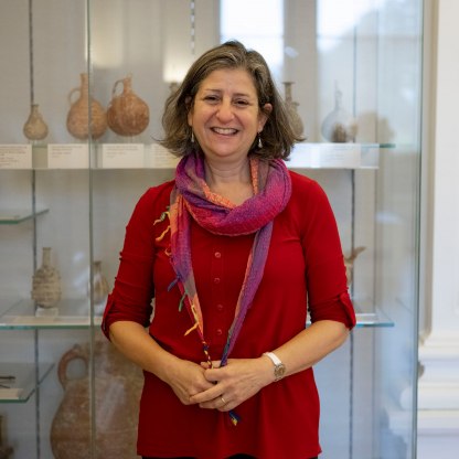 A profile image showing Susi Pancaldo in front of a case of objects. She's wearing a red top and a bright scarf